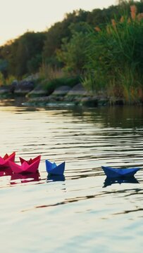 Different colored paper ships swim in a quick river surface on the natural background with green trees. Origami boats in bright colors on a lake at sunset Vertical video