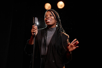 Waist up portrait of elegant Black woman performing on stage with microphone against dark background copy space