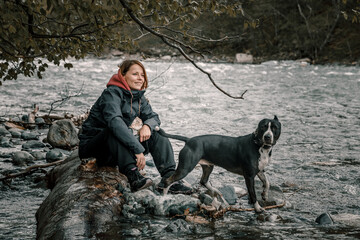 A beautiful girl and a dog are sitting on a log on the river bank. Beautiful dog. American Staffordshire Terrier. Cloudy weather.