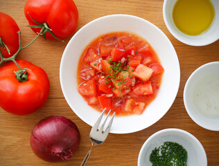 Top view of fresh tomato salad with red onion in white bowl and its ingredients on wooden table. Healthy vegetarian and vegan food taken from above.