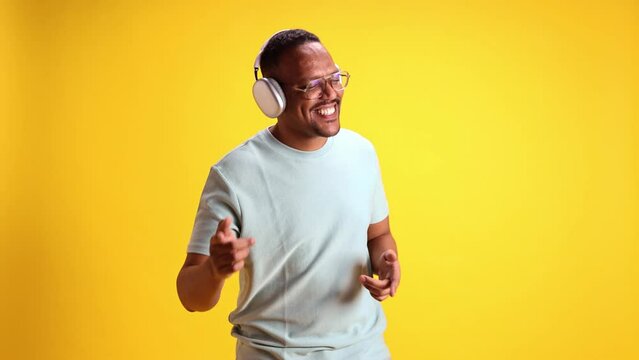 Cheerful, positive, relaxed African-American man in casual clothes listening to music in headphones and dancing against yellow studio background. Concept of human emotions, casual fashion, lifestyle