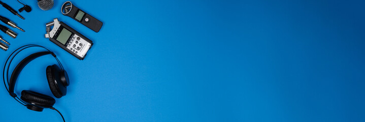 Banner, layout of accessories for recording and listening to sound: headphones, voice recorders, microphone, cables on blue background, top view. Concept of recording pure sound. Copy space.