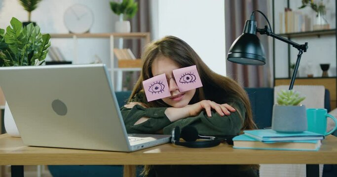 Beautiful tired woman falling asleep on the table near laptop and putting on glasses with funny stickers with drawn open eyes