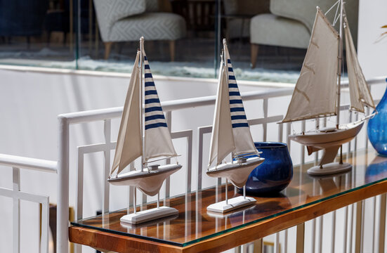 Nautical decor with model sailboats on a wooden table, interior.  Three model sailboats with beige sails, two of which have blue stripes. In the background a white railing and blue vases are visible