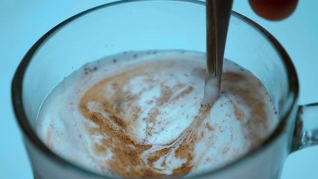 Hand with a spoon stirs white milk foam with the addition of cinnamon in a glass cup with cappuccino coffee. Closeup. Macro