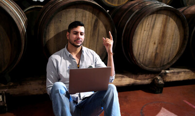Professional man winemaker working and inspecting wine quality in cellar with wooden barrel in...