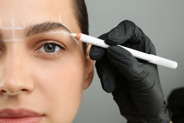 Beautician preparing young woman for procedure of permanent eyebrow makeup on grey background,...