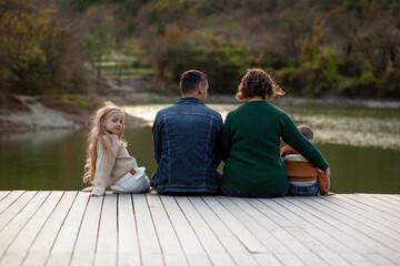 Back view of happy family of four people, mom, dad, daughter and son spend time outside the city on the lake together. Adult parents and children hugging and playing on a day off