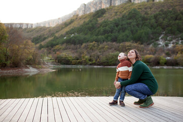 A happy mother holds a four-years-old child in his arms against the background of a lake, forest and mountain