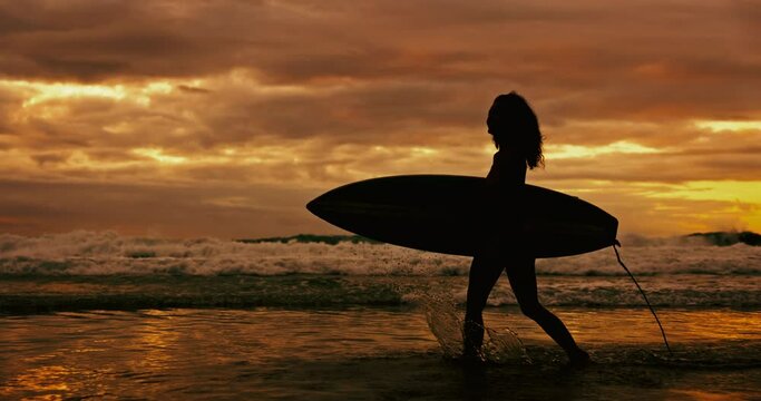 Sunset, silhouette and woman with a surfboard, beach and water with fitness and vacation. Person, girl and surfer with ocean and seaside with clouds and summer with wellness and nature with adventure