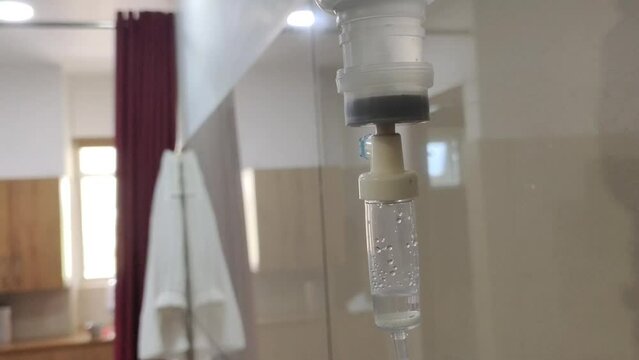 Hospital or clinic room with Intravenous IV drug infusion drip medicine liquid drops (sodium, glucose or saline solution) falling inside plastic tube. Closeup selective focus side view.
