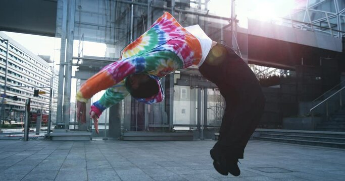 Super slow motion of young modern male break dancer in colorful urban street style wear is performing energetic choreography with backflip in the middle of urban city center streets.