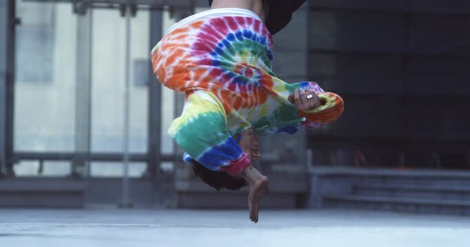 Super slow motion of young modern male break dancer in colorful urban street style wear is performing energetic choreography with backflip on stairs in urban city center.