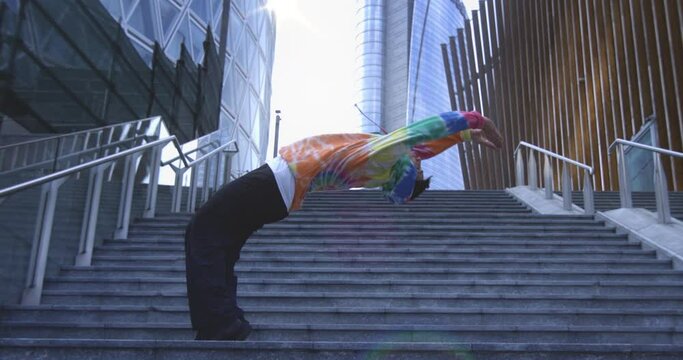 Super slow motion of young modern male break dancer in colorful urban street style wear is performing energetic choreography with backflip on stairs in urban city center.