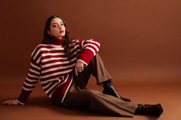Fashionable confident woman wearing stylish red striped woolen sweater, classic trousers, cowboy ankle boots, posing on brown background. Studio fashion portrait. Copy, empty, blank space for text
