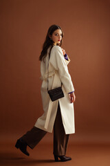 Fashionable confident woman wearing white coat, trousers, leather pointed toe boots, with small stylish shoulder bag, posing on brown background. Full-length studio fashion portrait