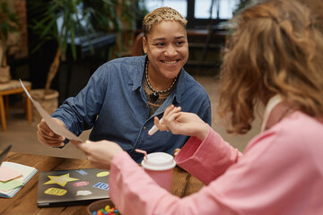 Portrait of smiling masc Black woman discussing project with colleague at meeting table and holding...