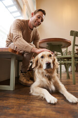 Vertical portrait of smiling handsome man with his dog, drinks coffee in pet-friendly cafe in city,...