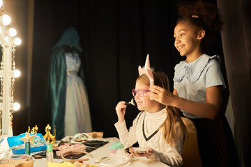 Side view portrait of two little girls doing makeup by mirror enjoying backstage preparations in...