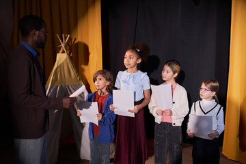 Diverse group of young children actors listening to drama teacher rehearsing school play on stage...