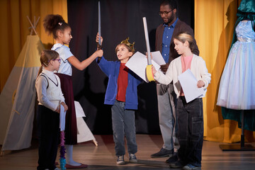 Full length portrait of children rehearsing school play on stage in theater with little boy prince...