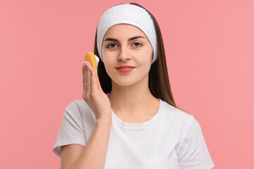 Young woman with headband washing her face using sponge on pink background