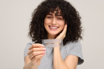 Young woman holding teeth whitening strips on light grey background, selective focus