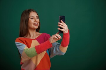 Looking and holding smartphone. Young woman is standing against green background in the studio
