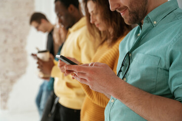 Side view. With smartphones in hands. Group of young people are standing against white background - obrazy, fototapety, plakaty