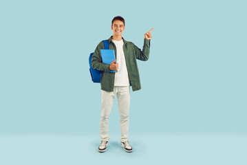 Portrait of a happy smiling young brunette boy holding notebook in hands with backpack on back pointing index finger to the side isolated on studio blue background. Education, knowledge concept.