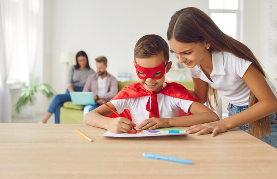 Happy children drawing at home. Little brother disguised in superhero cloak together with elder sister use power of imagination and draw colorful pictures on desk, with parents sitting in background