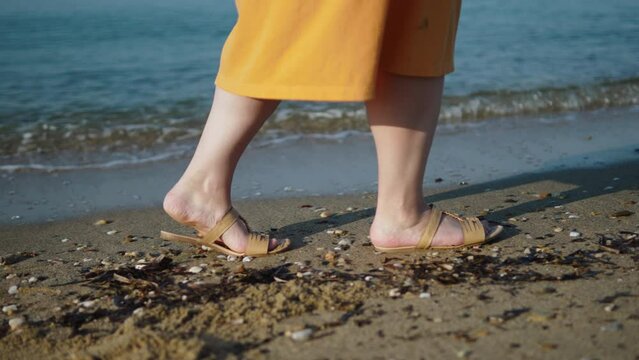 Unrecognizable overweight woman with flat feet walks in sandals along seashore, side view. Non-anatomical shoes with thin soles. Unhealthy shoes causing orthopedic problems.