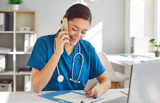 Friendly Young Woman Doctor, Nurse, Reception Specialist In Blue Scrubs With Stethoscope Sitting In Office Workplace, Making Phone Calls, Talking To Customers, Scheduling Appointments And Taking Notes