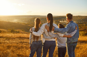 Back view of family of four hugging each other and looking into the distance enjoying sunset and beautiful nature standing in a row together. Young parents with children walking outdoors.