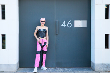 Young gay boy with pink hair and make-up and dressed in pink and black is leaning against a black garage door. The boy is wearing sunglasses and is doing different poses. Concept LGBTQ rights.