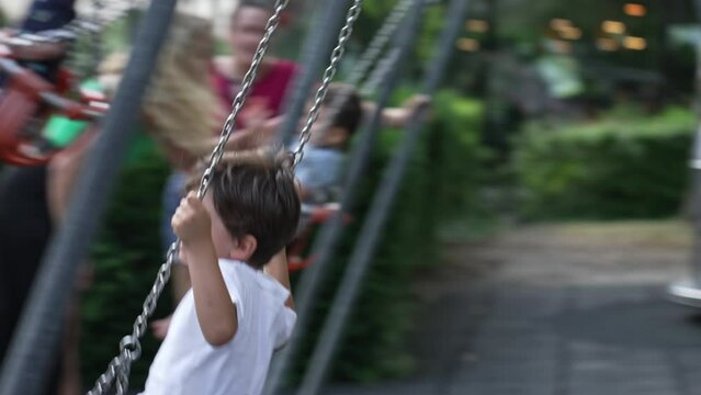 Mother Pushing Child In Playground Swing In The Midst Of Children. Parent And Son Bonding Moment, Mom Swings Young Boy In The Air During Weekend Activity At Park