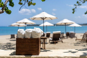 Roll up of white towels in basket on white table with copy space on blurred beach sunbeds and white...
