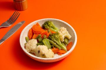 Steamed cauliflower, carrots, brocolli and vegetables inside a ceramic bowl ina orange background in front view