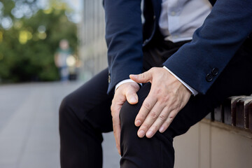 Close-up photo of young man's hands in a business suit holding the knee of the leg feeling severe...