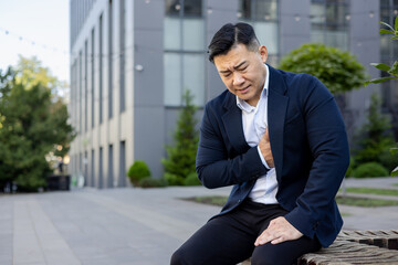 Young Asian male businessman sitting bent outside office building in suit, holding hand to chest and feeling severe pain in heart