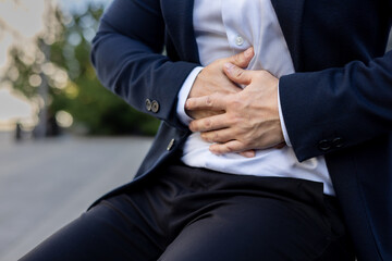 Hands of a young man in a business suit holding his stomach, sitting outside on a bench. Close-up photo