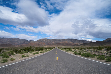 Road less traveled shown in the Mojave Desert in California, USA, in mid-March 2024. Road on a barren landscape with blue skies and puffy white clouds. Road forms a vanishing point.