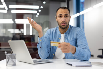 A man is sitting at a desk with a laptop and a credit card in his hand