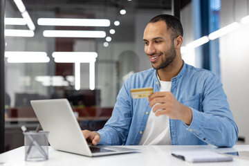 A man is smiling while holding a credit card in front of a laptop