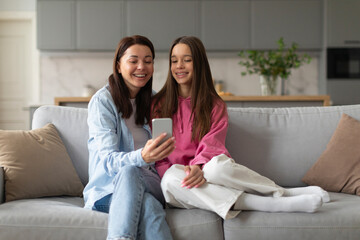 Happy mom with teen daughter using phone together at home, watching social media videos, doing online shopping on smartphone sitting on sofa together