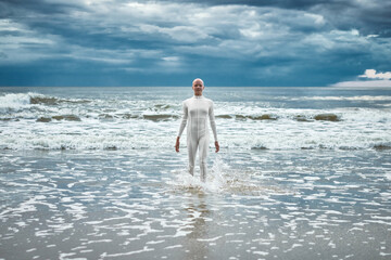 Brave hairless girl with alopecia in white futuristic suit comes out of cold sea on sandy beach,...