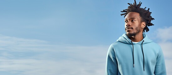A man with a beard in a blue hoodie is standing with one hand raised in front of an electric blue sky filled with fluffy clouds. He looks happy and ready to travel