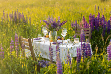 Romantic table decor for a loving couple on the blooming meadow with purple lupines. Two glasses of wine, flowers in a vase, silverware, fruits, wooden furniture and picnic basket. Sunset, golden hour