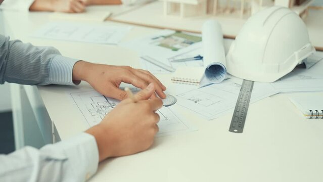 Close up of skilled architect engineer hand draws blueprint on meeting table with blueprint, architectural equipment, house model and safety helmet scatter around. Focus on hand. Immaculate.