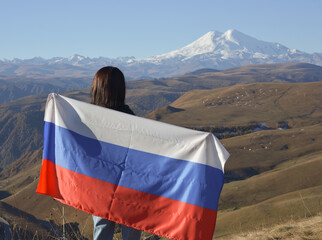 A young brunette woman stands against the backdrop of the snow-capped Mount Elbrus, looking at the beauty of the mountains, holding a Russian flag in her hands. Tricolor against the backdrop of snow